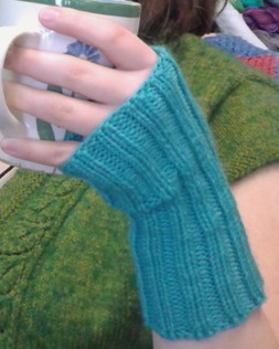 A lady's hand with short nails, wearing a handknit ribbed turqoise fingerless glove while holding a coffee mug