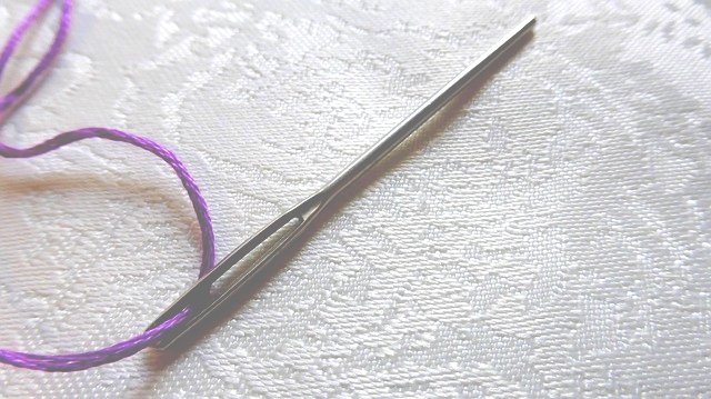 A silver-colored straight-tip darning needle threaded with purple yarn on a white damask fabric background